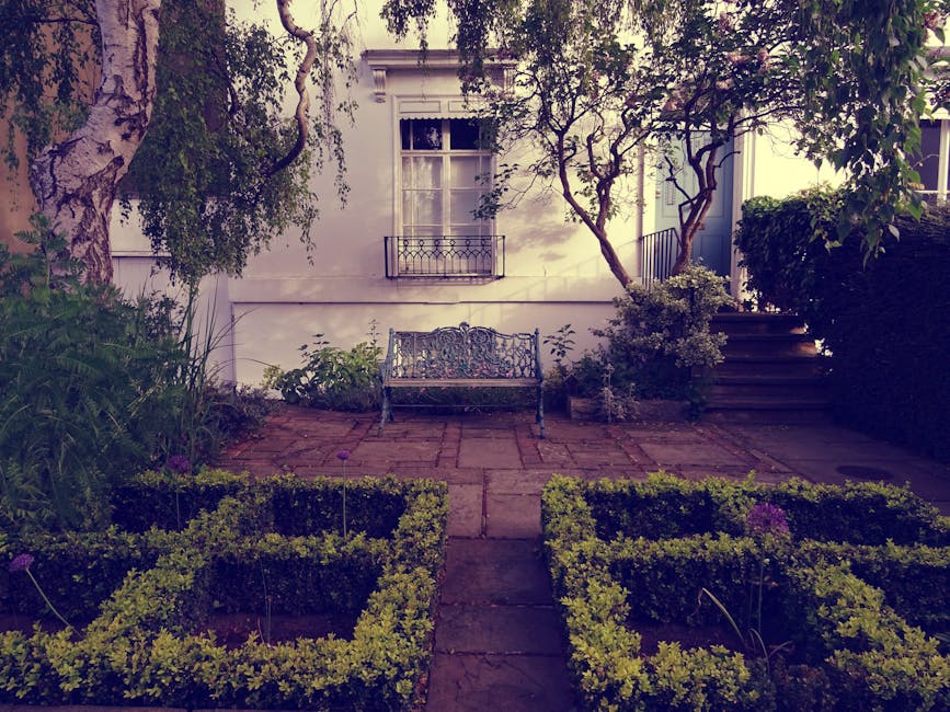 A small, well-maintained garden patio area featuring a central paved pathway leading to a decorative wrought iron bench positioned against a white exterior wall of a house with a window and balcony. T